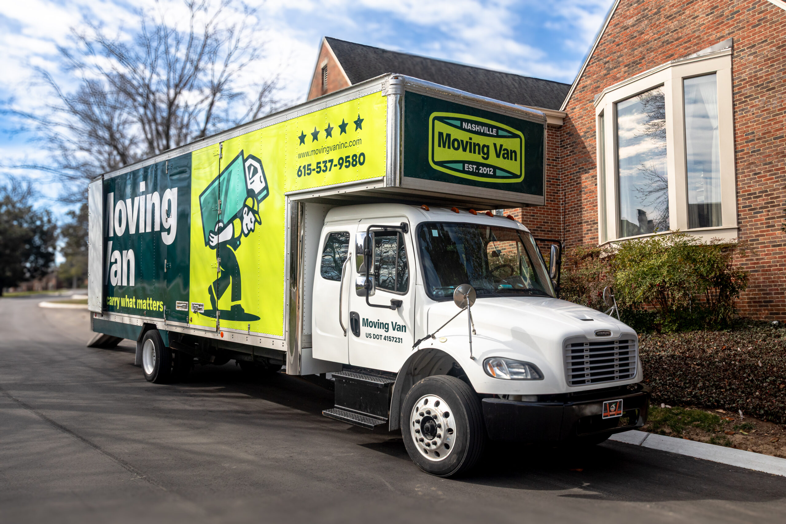 A large white moving truck with a green and yellow advertisement parked on a suburban street. The truck features cartoon movers, contact info, and a brick house nearby.