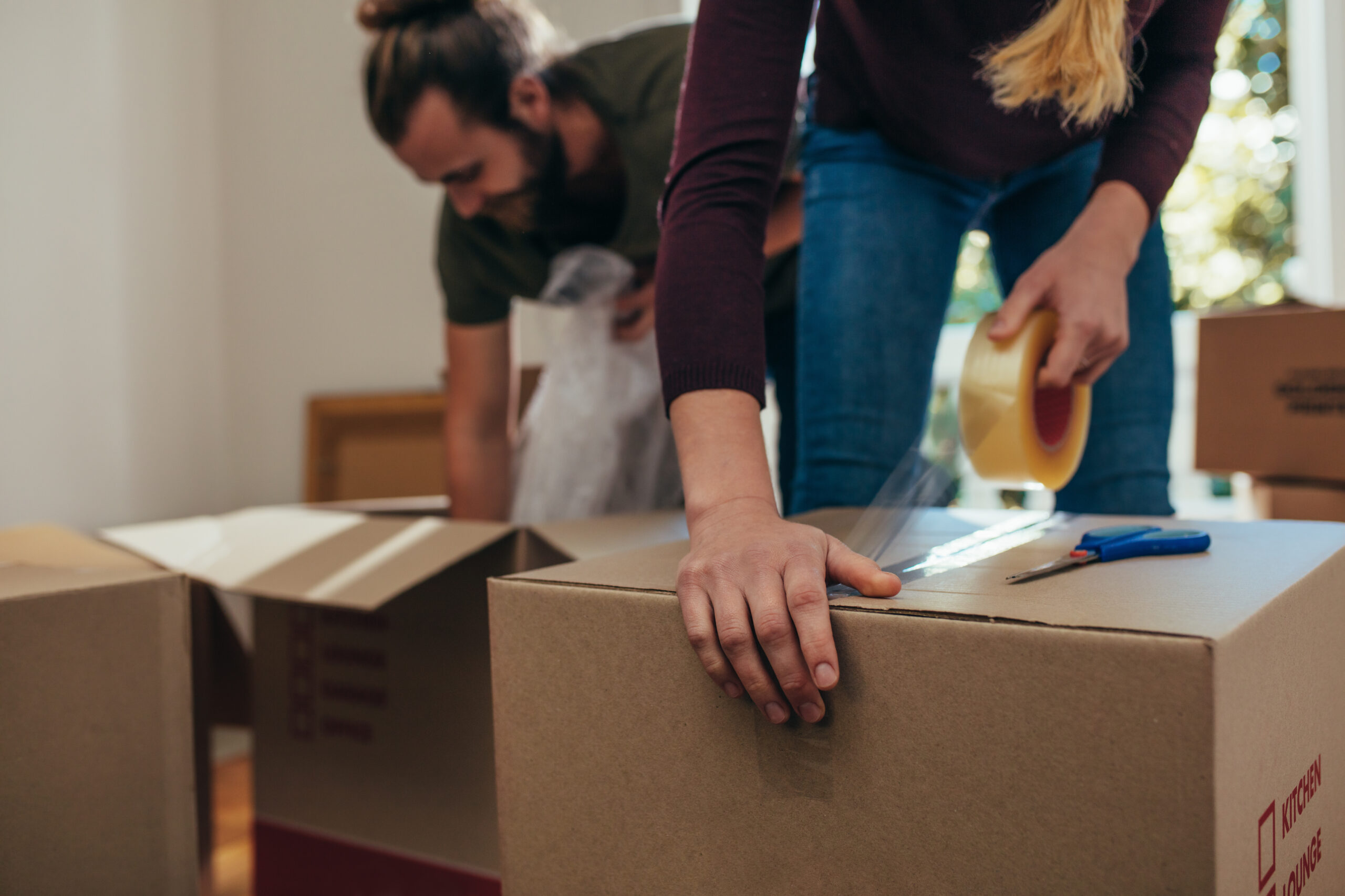 Couple using packing boxes to pack their household items. Man putting items in boxes while a woman applies adhesive tape on the boxes to seal them.